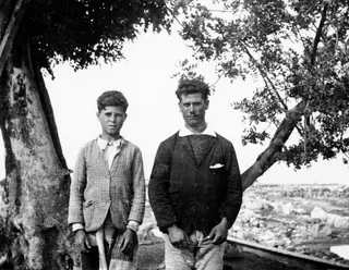 Two people stand side by side beside a large tree trunk, with low stone walls and rubble in the background at Vouni.