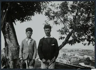 Two people standing beside a large tree at Vouni, with a stony field behind them