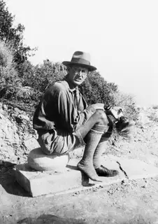 Einar Gjerstad seated on a worked stone block at Loures, Vouni, with bushes behind him.