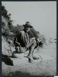Einar Gjerstad seated on cut stone blocks at Loures, Vouni, with bushes and a bank of soil behind him.