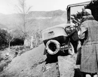 Car stuck at an angle on a grassy bank, with a man leaning into the driver's door and a person in a long coat watching.