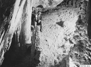 Two men on a rocky ledge inside a cave at Fylleri, Kythrea, with pale stone formations along the walls.