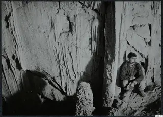 Man crouching in a cave at Kythrea, with rock walls streaked by vertical marks behind him