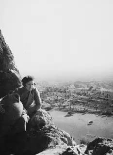Woman seated on a rocky ledge beside a large jar, with a wide valley and hills spread out below.