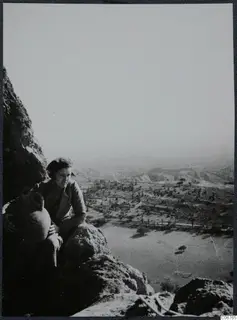 Woman seated on a rock ledge holding a large pottery vessel, with a valley and hills spreading out below