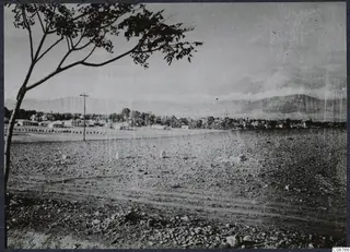 Low buildings and trees with mountains behind, seen across open stony ground at Nicosia.