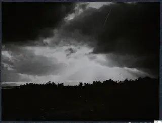 Sunbeams breaking through dark clouds above a line of trees on a flat plain, with faint mountains on the left horizon.