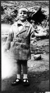 Young child standing on bare ground in a belted tweed coat, bow tie and headband, with a rocky bank behind