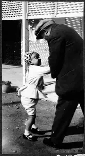 Man in a dark coat and flat cap leaning toward a small child in a patterned knit hat, on a dirt street in Nicosia.