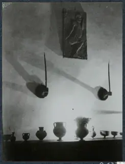 Row of vessels on a shelf in a hall, one holding a sprig of flowers, with a relief plaque and two wall-mounted objects above