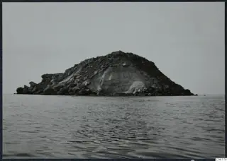 Rocky island rising from the sea under a pale sky, with a steep pale face on its right side