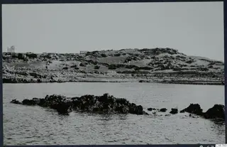 Rocky coastline with low scrubby hills, a small white building on the left, and dark rocks in the sea
