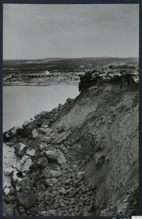 Eroded cliff edge with loose rocks at its foot, calm water on the left, and low hills and a small white building in the distance.