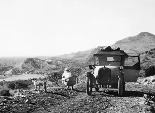 Boxy car on a stony track in hilly country, with a crouching person and a shaggy dog on a rise beside it