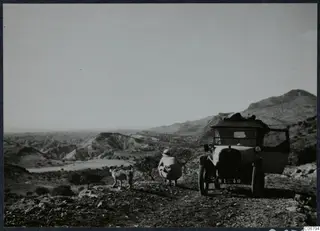 Landscape from the road to Hallefka, with a parked car, a crouching person and a dog on stony ground.