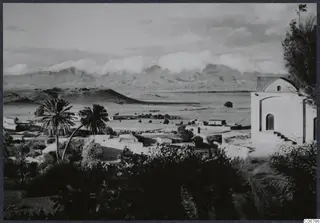 Village of flat-roofed houses and palm trees on a plain, with mountains behind and a whitewashed building at right