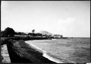Pebble shore and sea with low buildings, palm trees and a rounded hill, in the Kyrenia Mountains