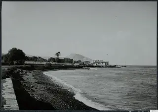 Pebble shore and sea with white buildings, palms and a rounded hill behind, in the Kyrenia Mountains area