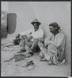 Two seated men against a whitewashed wall, with a small box and a pair of shoes on the ground beside them.