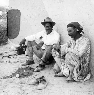 Two men sitting against a whitewashed wall at Ayios Iakovos, with a pair of shoes on the ground in front.