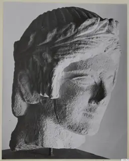 Limestone head from Mersinaki with carved hair in grooves and locks, shown on a dark stand