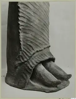 Lower part of a terracotta figure with a pleated skirt and two pointed feet, photographed close up against a plain background.