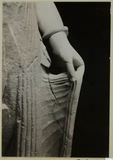 Close view of a limestone statue fragment showing a bare arm with a bracelet, the hand at the hip, and carved drapery folds.