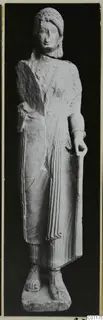 Limestone statue of a standing woman from Vouni, holding a long staff, with a broken left side and worn face.