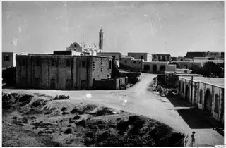 Town street with a large stone building, low houses and a tall slender tower, with one person at the lower right edge