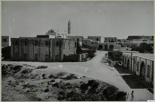 Street in Kyrenia with a large stone building, low houses, shrubs and one person standing at lower right.