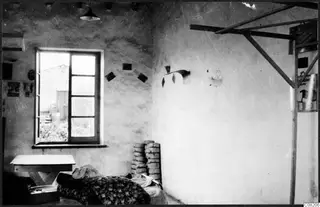 Interior of a plastered room with a window, a low wooden table and a bed covered by a floral blanket.