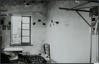 Interior of a plastered room with a window, a wooden table and a bed covered by a floral blanket.