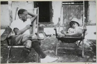 A man and a small child seated in wooden deck chairs outside a whitewashed building at Machairas.