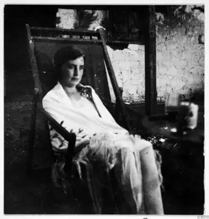 Woman sitting in a wooden deck chair wearing a light shawl, in front of a plastered wall.