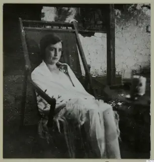 Woman seated in a wooden deck chair, wearing a light shawl, in front of a plastered wall.