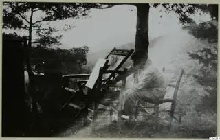 A man seated on a wooden folding chair under a tree, working beside a board on a stand, with hills behind