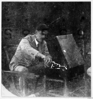Man seated in a chair at Machairas, handling a dark metal apparatus with bright fittings beside a wooden box.