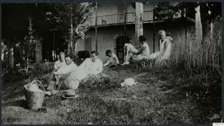 Group of people sitting on grass in front of a two-storey house with a balcony, with a basket and two hats on the lawn.
