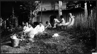 People sitting on a grassy slope in front of a two-storey house, with a wicker basket and hats in the foreground.
