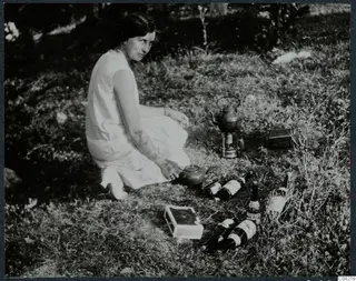 A woman sitting in grass among bottles, with a kettle on a small portable stove beside her