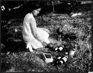 Woman sitting in grass beside glass bottles, a teapot and a small metal portable stove with a kettle