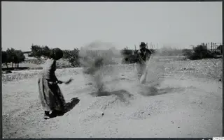 Two people working a patch of loose gravel in a dusty field, with bushes and trees behind them