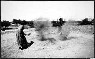 Blurred photo of a woman in a long dress and a man in a hat beside a dust cloud on dry gravelly ground, with trees behind.