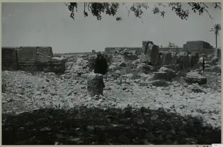 Man in a hat and dark coat standing among stone ruins, with a cylindrical column drum in the foreground