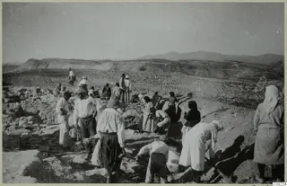 Workers in light clothing digging at an excavation among stone walls, with hills and cultivated fields behind