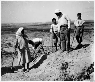 Five people with shovels and a spade at an excavation cut into dry ground, with low hills behind