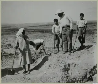 Five people with digging tools at the edge of an excavation, including a man in a pith helmet, with open hills behind.