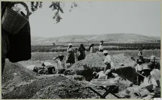 Workers digging an excavation trench beside spoil heaps, with a woven basket at top left and a folding stool in the foreground.
