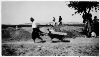Man in a light shirt and hat pushing a wheelbarrow along a dirt track past earth mounds, with people and hills behind.