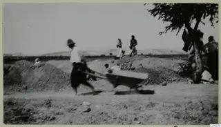 A blurred man pushes a wheelbarrow past earth heaps, with women standing nearby and low hills behind